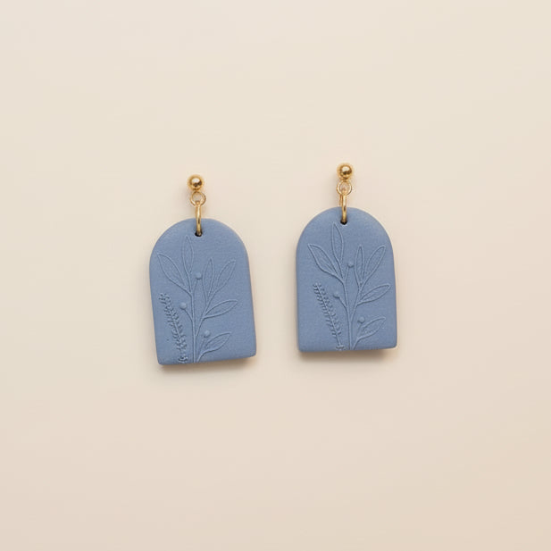 A pair of blue arch-shaped earrings with a wildflower texture, displayed on a white surface with dried flowers around.