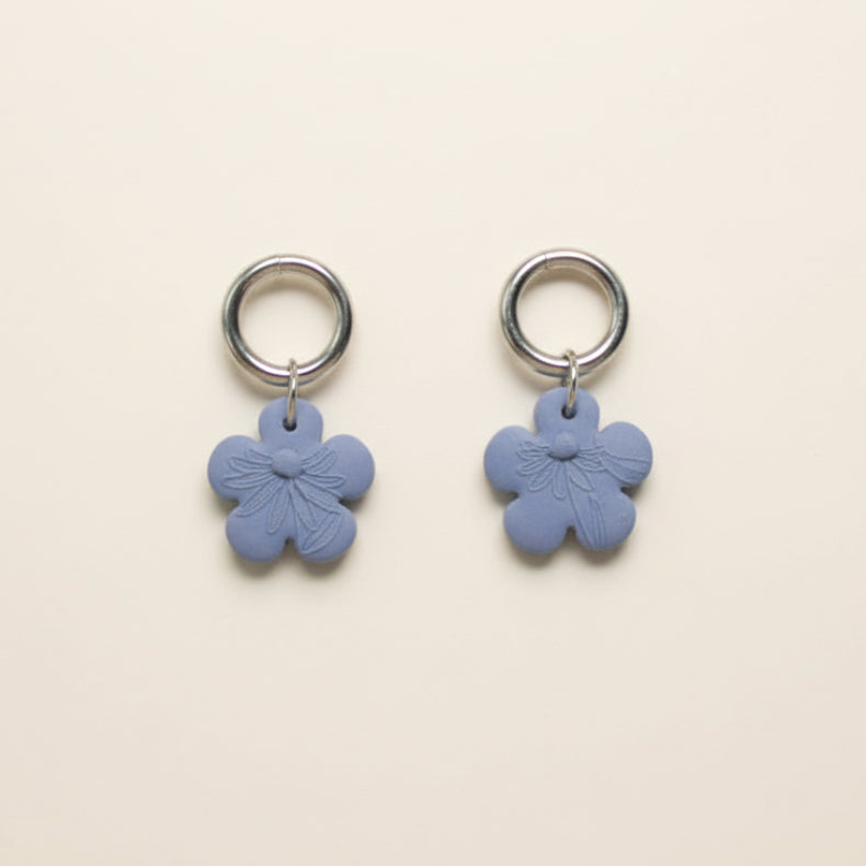 A pair of blue flower-shaped earrings with ring stud toppers, displayed on a white surface with dried flowers around.