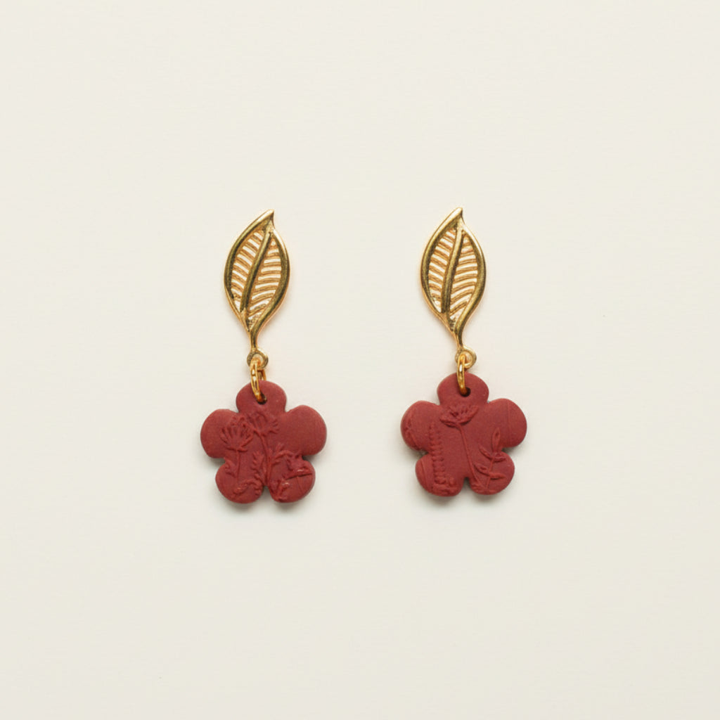 A pair of red flower-shaped earrings with gold leaf stud toppers, displayed against a white background with dried flowers around it.