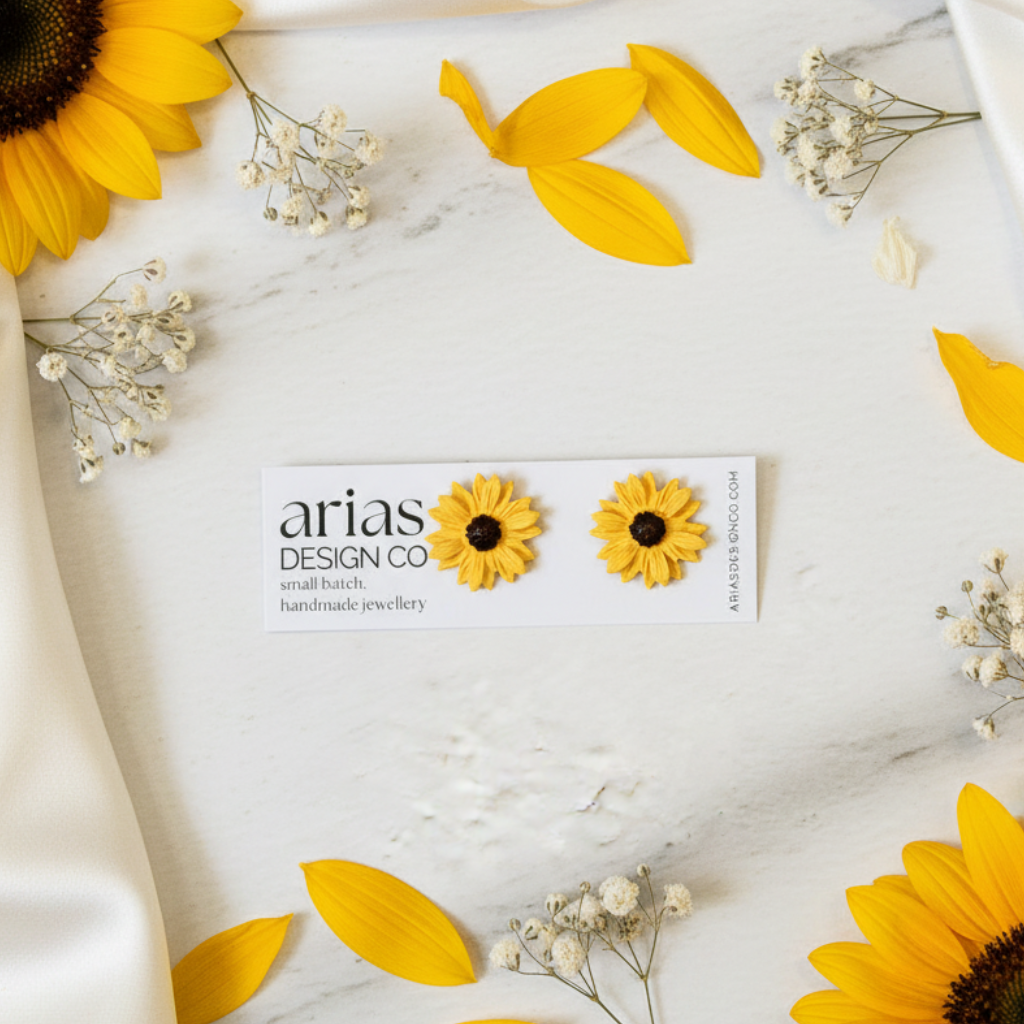 Sunflower stud earrings with 'arias design co' packaging on a marble surface with sunflowers and leaves.