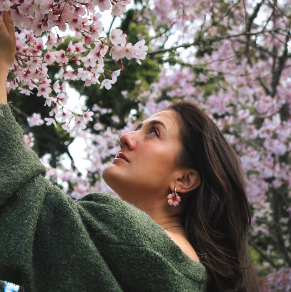 A woman in a textured green sweater looks up at pink cherry blossoms, wearing pink flower-shaped earrings. The background is filled with blooming cherry blossoms and soft green foliage.