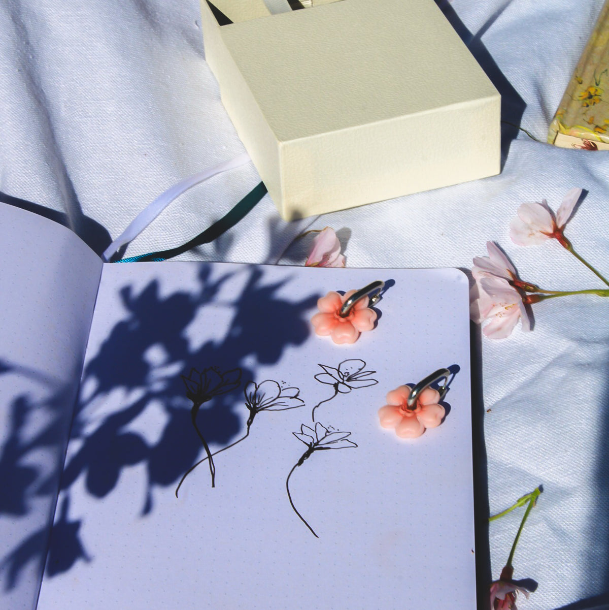 cherry blossom huggies earrings in silver on an open sketchbook with a gift box and flower shadows